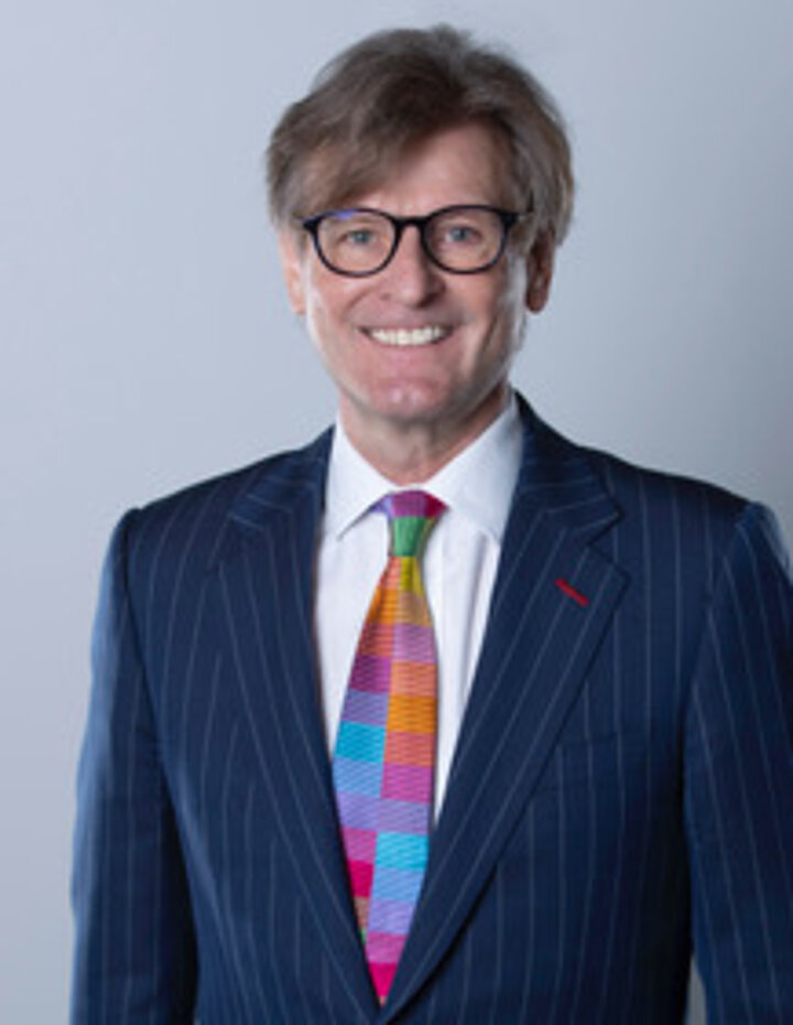 Portrait photo of a man in a blue suit. He wears a colorful tie and black glasses.